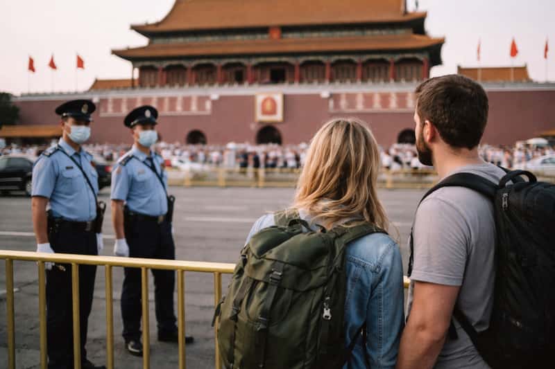 American tourist holding passport and map at Beijing train station, studying departure board with concerned expression